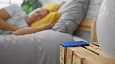 Rahat bir şekilde rahat bir yatağa kurulmuş, yakışıklı orta yaşlı bir adam yatak odasında huzur içinde uzanıyor, dinlendirici bir uykuya dalmaya hazırlanıyor, dinlendirici bir oda zeminine karşı, telefonla.