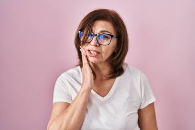 Orta yaşlı İspanyol kadın pembe arka planda durup diş ağrısı ya da diş ağrısı yüzünden elleriyle dudaklarına dokunuyor. dişçi 