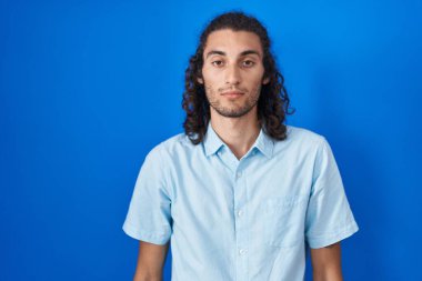 Mavi arka planda duran Latin asıllı genç adam yüzünde ciddi bir ifadeyle rahatlamış. Basit ve doğal bir şekilde kameraya bakıyorsun.. 