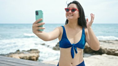 Bikini giyen genç Çinli kadın turist deniz kenarında video görüşmesi yapıyor.