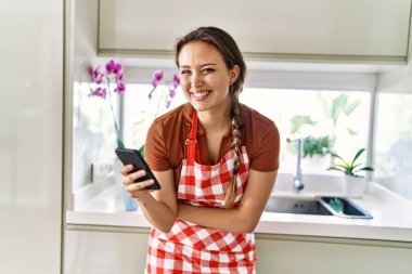 Mutfakta akıllı telefon kullanan önlük giyen genç ve güzel bir İspanyol kadın.