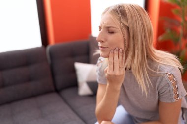 Diş ağrısı çeken genç sarışın kadın evdeki kanepede oturuyor.