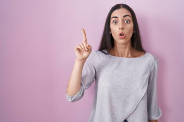 Young brunette woman standing over pink background pointing finger up with successful idea. exited and happy. number one. 