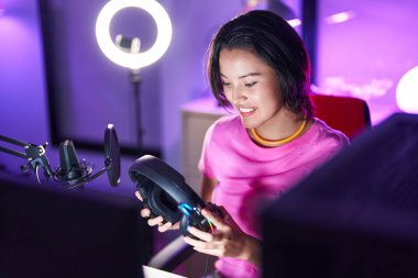 Young hispanic woman streamer smiling confident holding headphones at gaming room
