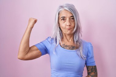 Pembe arka planda duran orta yaşlı bir kadın güçlü kol kası gösteren, kendinden emin ve gururlu biri. 