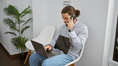 Genç İspanyol adam dizüstü bilgisayar kullanıyor. Akıllı telefonla konuşuyor. Bekleme odasında sandalyede oturuyor.