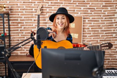 Müzik stüdyosunda klasik gitar çalan beyaz bir kadın mutlu mesut elini ve parmağını göstererek gülümsüyor. 