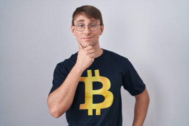 Bitcoin t-shirt giyen beyaz sarışın adam kameraya kendinden emin bakıyor kollarını kavuşturmuş ve çenesini kaldırmış gülümsüyor. Olumlu düşünmek. 