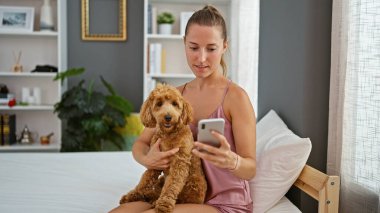 Köpekli beyaz bir kadın yatak odasındaki yatakta akıllı telefon kullanıyor.