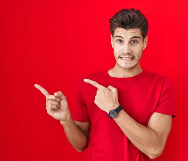 Young hispanic man standing over red background pointing aside worried and nervous with both hands, concerned and surprised expression 