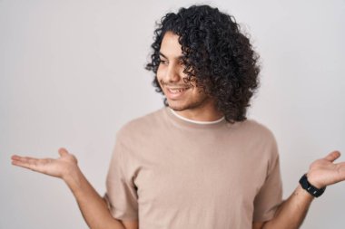 Beyaz arka planda duran kıvırcık saçlı İspanyol adam her iki elinde açık avuçlar, sunum ve reklam karşılaştırma ve denge gösteren 