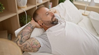 Yakışıklı genç bir adam rahat bir yatakta uzanıyor, yatak odasında dinleniyor, ellerini başının üstüne koyuyor.