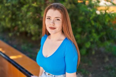 Genç, kızıl saçlı, kendine güvenen bir kadın parkta bankta oturuyor.