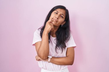 Pembe arka planda, elleri çenesinde soru soran genç İspanyol kadın, düşünceli bir ifade. Düşünceli bir yüzle gülümsüyor. şüphe kavramı. 