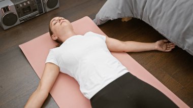 Uyanmış ve sakince nefes alıyor, güzel sarışın bir kadın yatakta uzanıyor, sabahını yoga egzersizlerini minderinde yapmaya adamış, yatak odasında denge ve sağlığa konsantre oluyor..