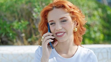 Genç kızıl saçlı kadın parkta gülümseyen akıllı telefondan konuşuyor.