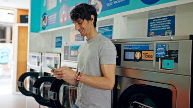 Çamaşır makinesine yaslanmış, çamaşır makinesinde gülümseyen akıllı telefon kullanan genç bir İspanyol.