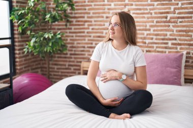 Genç hamile kadın gülümsüyor. Yatak odasında karnına dokunuyor.