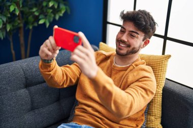 Genç ispanyol adam evdeki kanepede oturarak akıllı telefonuyla selfie çekiyor.