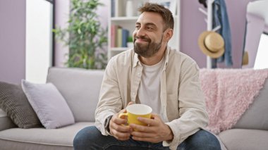 Neşeli bir sabah birası, genç, sakallı İspanyol adam kendine güven ve mutluluk saçıyor, gülümsüyor, elinde bir kahve fincanı tutuyor ve şık dairesinde kanepede dinleniyor..