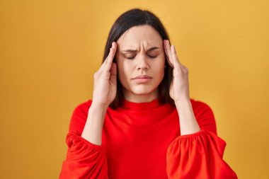 Young hispanic woman standing over yellow background with hand on head for pain in head because stress. suffering migraine. 