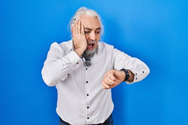 Mavi arka planda gri saçlı, orta yaşlı bir adam. Saate bakıyor. Endişeli, geç kalmaktan korkuyor. 