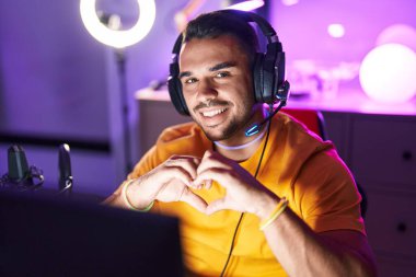 Young hispanic man streamer smiling confident doing heart symbol with hands at gaming room