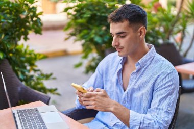 Young hispanic man using smartphone sitting on table at coffee shop terrace