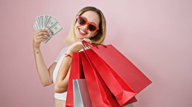 Genç sarışın kadın alışveriş torbaları ve dolarları soyutlanmış pembe arka planda tutuyor.