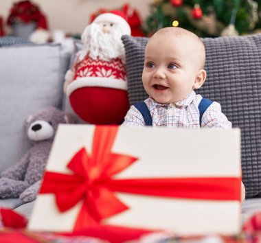 Evdeki Noel ağacının yanındaki koltukta oturan sevimli beyaz bebek hediyesini tutuyor.