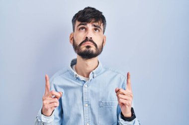 Mavi arka planda dikilen sakallı genç İspanyol adam üzgün ve üzgün görünüyor, parmaklarıyla yönü gösteriyor, mutsuz ve depresif.. 