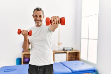 Orta yaşlı, gri saçlı bir hasta rehabilitasyon merkezinde dambıl kullanıyor.