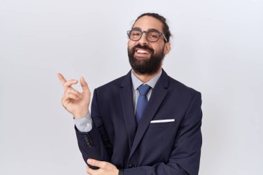 Hispanic man with beard wearing suit and tie with a big smile on face, pointing with hand and finger to the side looking at the camera. 