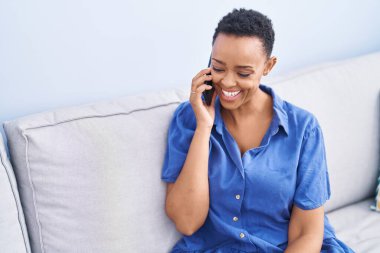 Afro-Amerikalı kadın akıllı telefonda konuşuyor. Evdeki kanepede oturuyor.