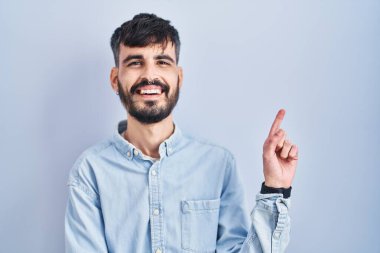 Mavi arka planda duran sakallı, yüzünde kocaman bir gülümseme olan, eli kameraya bakan genç İspanyol bir adam.. 