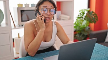 Genç, güzel, İspanyol bir kadın. Laptop kullanıyor. Akıllı telefondan konuşuyor.