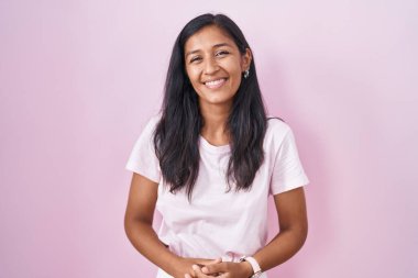 Young hispanic woman standing over pink background with hands together and crossed fingers smiling relaxed and cheerful. success and optimistic 