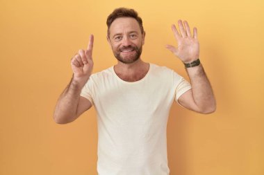 Sarı arka planda sakallı orta yaşlı bir adam 6 numaralı parmaklarıyla işaret ederken kendinden emin ve mutlu gülümsüyor.. 