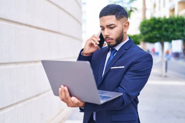 Young latin man business worker using laptop talking on smartphone at street