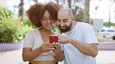 Kendine güvenen, mutlu ırklar arası bir çift birlikte gülümsüyor, akıllı telefonlarıyla güneşli parkta duruyor, açık havada sevgiyi ve kahkahaları zahmetsizce paylaşıyorlar.