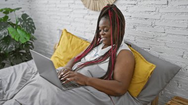 Güzel, kendine güvenen Afro-Amerikan kadın, ışıl ışıl gülümseyen, dizüstü bilgisayarla eğlenen, rahat bir yatakta, kapalı bir odada, yatak odasında, sabahları