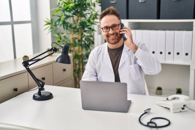 Kafkasyalı genç doktor laptopunu kullanarak klinikte akıllı telefondan konuşuyor.