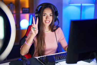 Genç İspanyol kadın video oyunları oynuyor. İki numarayı gösterip işaret ediyor. Kendinden emin ve mutlu bir şekilde gülümsüyor.. 