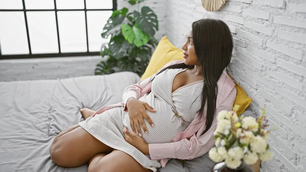 Young pregnant woman seeking tranquility while gently touching her belly, resting on her bed in a cozy bedroom atmosphere, reflecting on the joys of upcoming motherhood