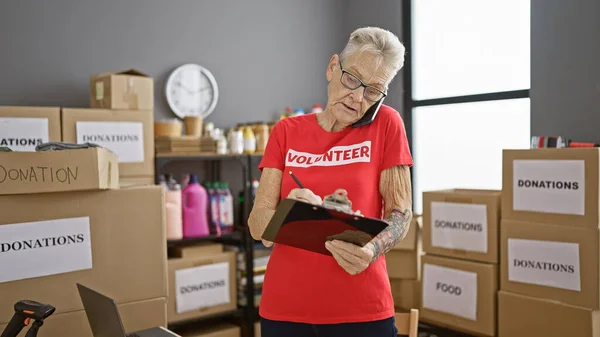 Senior grey-haired woman volunteer talking on smartphone taking notes at charity center