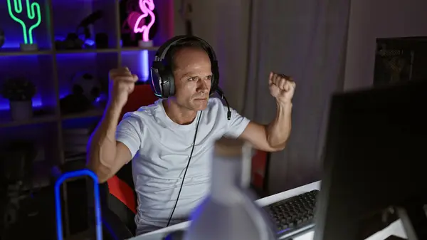 陽気な中年男性は 手軽にリラックスし 勝利の夜のゲームをストリーミング テクノロジーに吸収されたダークゲームルームでビデオゲームをプレイするヒスパニックストリーマー — ストック写真