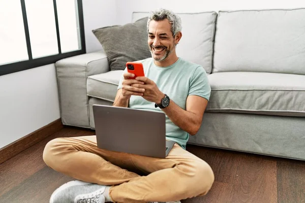Middle age grey-haired man using smartphone and laptop sitting on floor at home