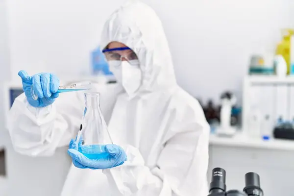 Young blonde woman scientist wearing security uniform pouring liquid on test tube at laboratory