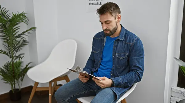 stock image Young hispanic man sitting on chair reading document on clipboard at waiting room