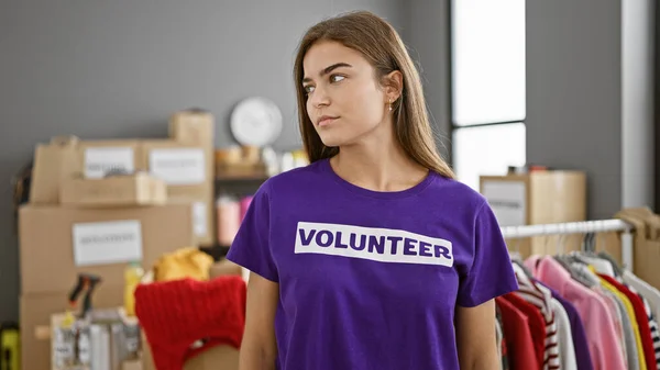At the heart of community service, young, beautiful hispanic woman volunteers her time, standing in uniform with a serious face indoors at charity center, a warehouse filled with donations.